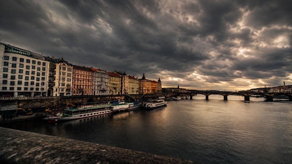 Prague Vltava riverfront embankment with historic buildings, river cruise boats, and bridge under dramatic sunset clouds